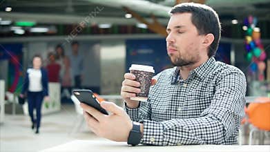 A man with a concentrated look typing sms on a mobile phone, in his hand coffee.