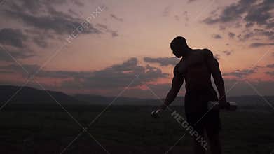 Silhouette of african american muscular body builder lifting dumbbells against the pink sunset sky background. Outdoor