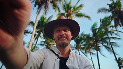A young man in a hat with moustache among coconut palms keeps the camera or phone in your hands, shoots himself and