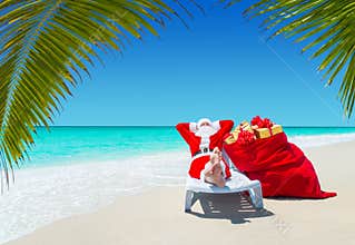 Santa Claus with Christmas sack full of gifts relax on sunlounger at tropical palm beach.