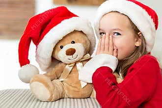 Cute young girl wearing santa hat whispering a secret to her teddy bear christmas present toy. Cheeky kid with teddy bear.