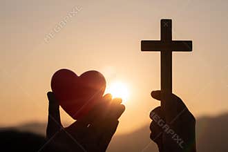 Silhouette of woman hands praying with cross and holding a red heart ball in nature sunrise background, Crucifix, Symbol of