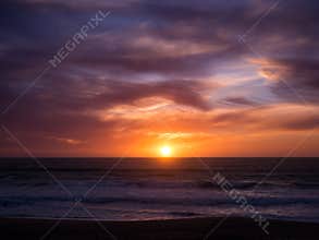 Sun on horizon at sunset with blue orange gradient sky and dark dramatic clouds over ocean