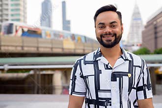 Happy young bearded Indian man smiling against view of train at sky train station