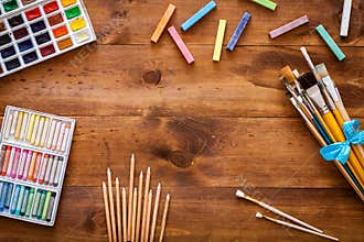 Creative art work accessories tools supplies set on messy desk, paint brushes, paintbox watercolors crayons pencils on brown