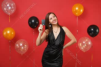 Dreamy woman in little black dress celebrating looking up, holding credit card thinking how to spend money on bright red