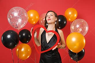Dreamy young woman in little black dress celebrating, looking up, holding big red wooden heart on red background air