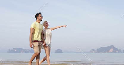 Couple Walking On Beach Holding Hands Talking, Young Man And Woman Point Finger, Tourists On Vacation