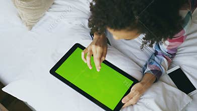 Top view of curly mixed race woman lying in bed at home using electronic tablet with green screen