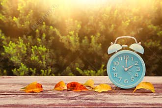 Image of autumn Time Change. Fall back concept. Dry leaves and vintage alarm Clock on wooden table outdoors at afternoon