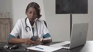 Black female doctor sits at an office desk looking to camera, african american nurse work on laptop in cabinet, modern