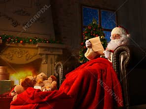 Portrait of happy Santa Claus sitting at his room at home near Christmas tree and reading Christmas letter or wish list.