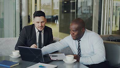 Two multi ethnic businessmen looking at laptop computer and laughing in glassy cafe. Business collaegues having fun and