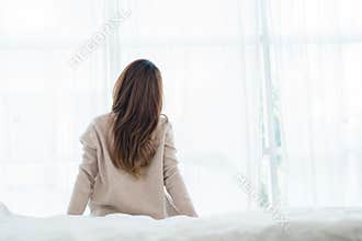Back view of happy beautiful young Asian woman waking up in morning, sitting on bed, stretching in cozy bedroom.