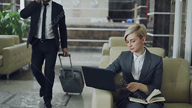 Blonde businesswoman sitting in armchair with notepad and laptop computer while businessman with luggage walking through