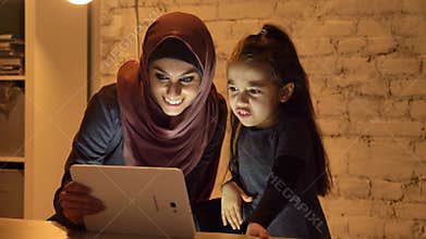 A young mother in a hijab sits with her little daughter on the couch in the evening and uses a tablet, watch a funny