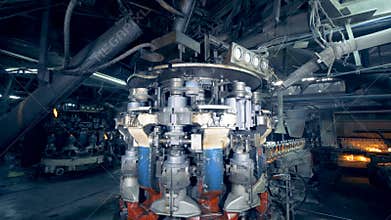 A metal machine rotates at a plant, while bottles move on an assembly line behind. 4K.