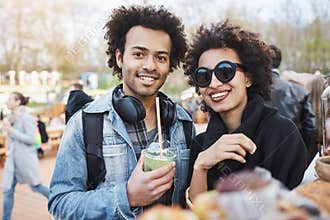 Portrait of happy cute dark-skinned couple with afro hairstyle, strolling on food festival, tasting and drinking
