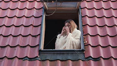 Woman enjoying morning looking out the roof window of house and drinks coffee