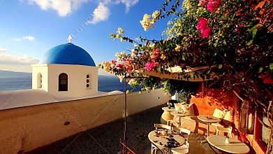 Blue church dome with wind playing with colorful flower shrub on a terrace of traditional cafeteria in typical greek