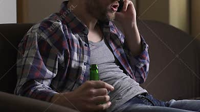 Angry addicted man sitting on couch, talking on cellphone and drinking beer