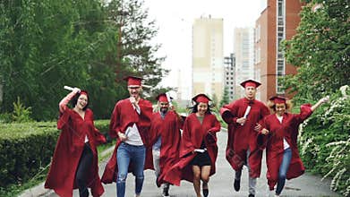Excited graduating students running along road on campus holding diplomas wearing graduation clothes gowns and hats