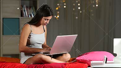 Serious teen writing in a laptop sitting on a bed at home