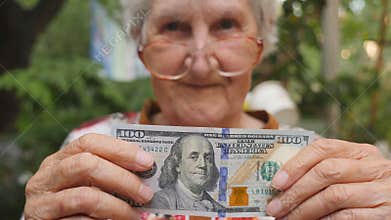 Old woman in eyeglasses showing one hundred dollar bill into camera outdoor. Happy grandmother holding foreign currency