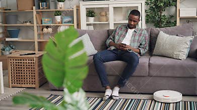 Carefree African American man is using robotic vacuum cleaner sitting on sofa and holding smartphone enjoying