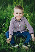 Portrait of stylish blonde little boy sitting in park among grass.