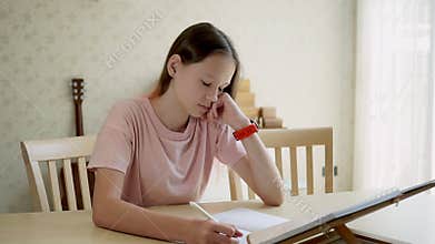 Cute teen girl girl looks at a book and writes in a notebook then looks at her watch. homeschooling concept. Living room in backgr