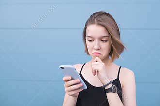 Pretty girl with a sad face looks at a smartphone on the background of a blue pastel wall