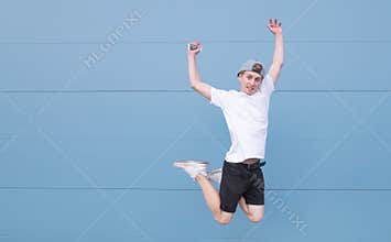 Emotional young man in a white T-shirt jumps against the background of a blue wall and looks at the camera