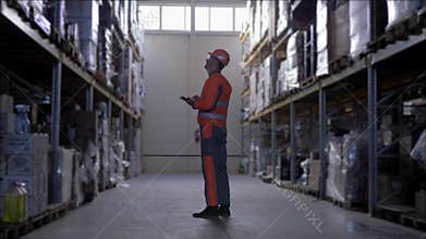 Warehouse worker with glasses in hard helmet and uniform makes notes on tablet during carries out an audit