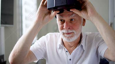 Good-looking handsome senior man in white takes off VR 360 glasses at home. Active elderly people