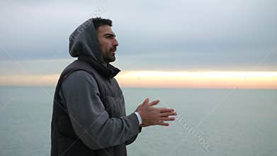 Man is warming up his hands by breathing at the cold morning seaside. Morning sunrise over the sea.