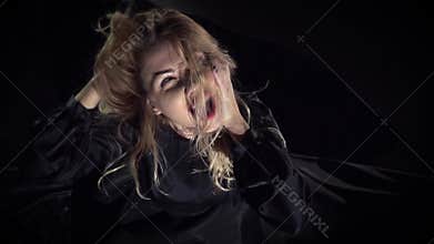 A blond witch with red lips sits on the floor, ruffling her hair and yelling fiercely, slow motion