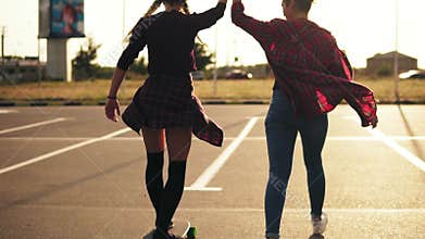 Back view of young attractive hipster girl being taught skateboarding by a friend who is supporting her holding her hand