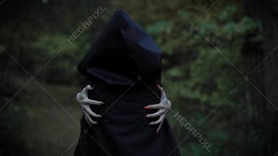 A woman in a cloak with a hood embraces herself behind her hands with long nails, like a witch, a mystical lady walks