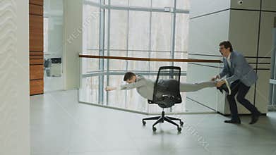 Slow motion pan shot of two funny businessmen riding office chair while having fun in lobby of modern business center