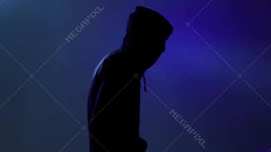 An impersonal person hunched over and turns to the camera. Abstract man in black clothes with a hood passes on a blue luminous bac