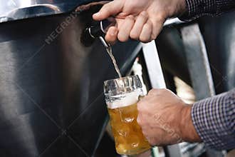 Strong man`s hands pour beer in tumbler from beer tap at craft brewery.
