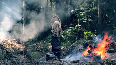 A young woman stand near fire and raises her hands and lowers them