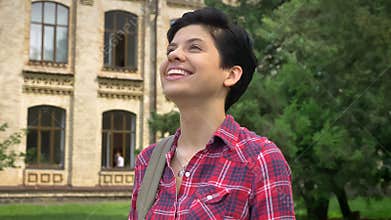 Happy young college student with black short hair smiling and standing in park near university
