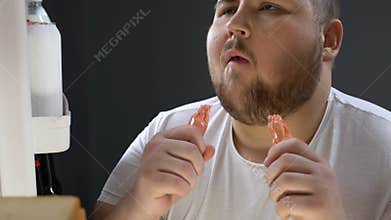 Greedy obese man eating sausages plastic cover near fridge at night, consumerism
