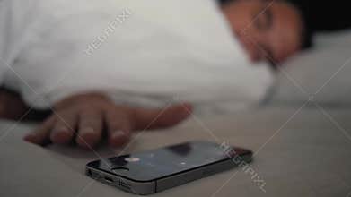 Young man sleeping on the bed with an alarm clock on the mobile phone display. Modern technologies