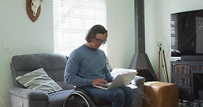 Happy caucasian disabled man in wheelchair using laptop in living room