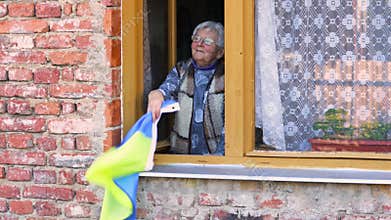 Ukrainian grandmother waving with Ukraine flag from the window, praying peace, Russian aggression to Ukraine at 2022