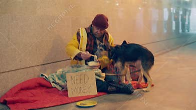 A vagrant man is feeding his dog while sitting on the ground