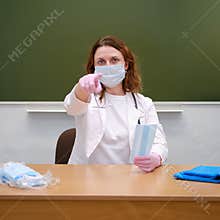 School nurse in a medical mask sits at her desk and points her index finger at you, copy space